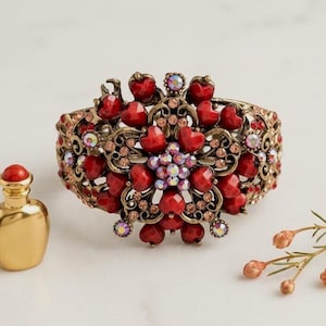 Può includere: Un bracciale a fascia larga con una base in filigrana di metallo color ottone anticato, decorato con numerose perline rosse a forma di cuore e piccoli cristalli iridescenti disposti in un motivo floreale al centro e sui lati.