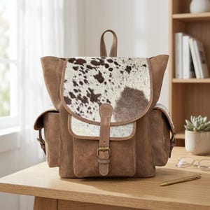 May include: A brown backpack featuring a cowhide print flap with dark brown and white patches. The bag is constructed from a smooth brown material with side pockets, a top handle, and a central vertical strap secured by a brass-toned metal buckle.
