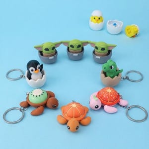 May include: A collection of small animal-themed keychains and figurines. The set includes three green alien-like figures in grey pots, a penguin in a white egg, a green dinosaur in a brown egg, and three turtles with fruit-patterned shells.