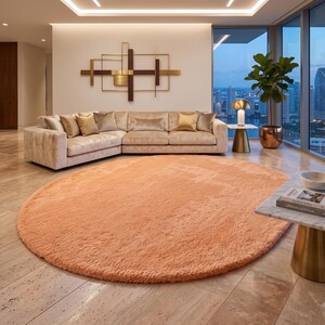 May include: A large, round, peach-colored area rug with a thick, plush, shaggy texture. The rug is positioned on a light-colored stone floor in front of a beige sectional sofa, providing a soft, textured contrast to the smooth, hard flooring.