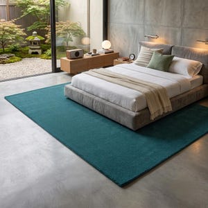 A solid teal area rug lays flat on a light grey stone floor in a modern, minimalist room. The vibrant blue-green hand-tufted rug anchors the space next to a natural wood side table with smooth black stones and a candle. A sleek beige daybed sits nearby, looking out onto a serene Japanese zen garden through large glass doors. IMG_20260727_232441.jpg displays its rich, plush texture beautifully.