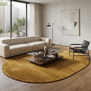 May include: A large oval mustard yellow area rug placed on a wooden floor in a living room. The rug sits beneath a two-tiered coffee table with a glass top and wooden base, a beige sofa, and a black armchair with a metal frame and leather seat.