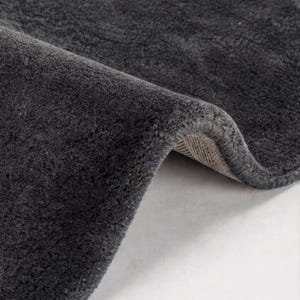 May include: A dark charcoal gray rug with a dense, plush pile texture. The rug is folded over to reveal a light beige, woven fabric backing along the curved edge, showcasing the thickness and construction of the material in a close-up view.
