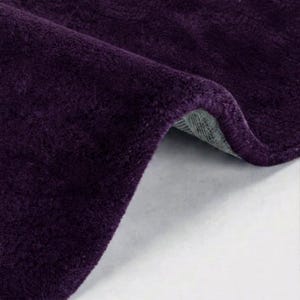 May include: A deep purple rug with a soft, plush texture. The rug is folded over to reveal a light gray, woven backing material. The surface of the rug is uniform in color and features a dense, short pile construction throughout the entire piece.