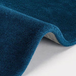 May include: A deep teal area rug with a dense, plush pile texture. The rug is folded over to reveal a light gray, woven backing material. The surface of the rug features a uniform, solid color with a soft, velvety appearance and short fibers.