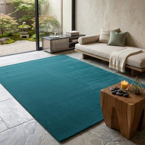 A solid teal area rug lays flat on a light grey stone floor in a modern, minimalist room. The vibrant blue-green hand-tufted rug anchors the space next to a natural wood side table with smooth black stones and a candle. A sleek beige daybed sits nearby, looking out onto a serene Japanese zen garden through large glass doors. IMG_20260727_232441.jpg displays its rich, plush texture beautifully.
