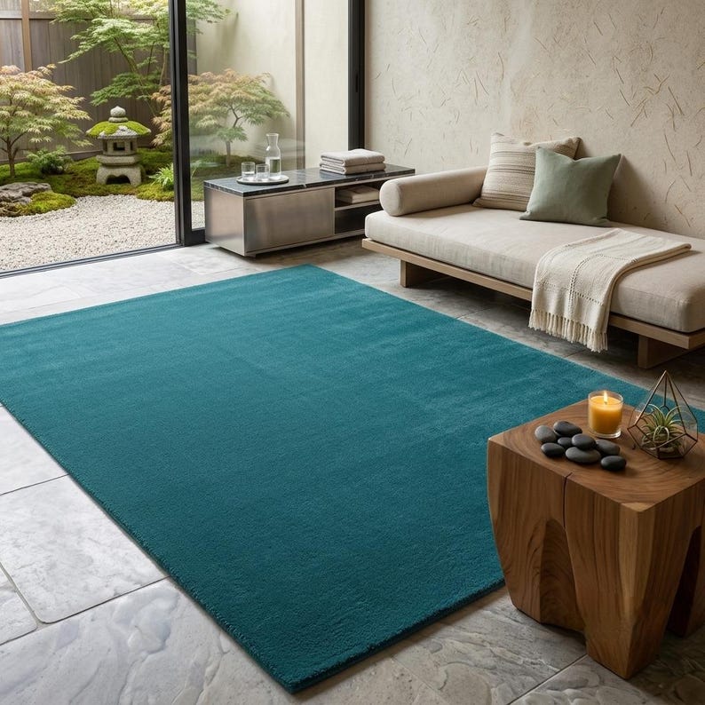 A solid teal area rug lays flat on a light grey stone floor in a modern, minimalist room. The vibrant blue-green hand-tufted rug anchors the space next to a natural wood side table with smooth black stones and a candle. A sleek beige daybed sits nearby, looking out onto a serene Japanese zen garden through large glass doors. IMG_20260727_232441.jpg displays its rich, plush texture beautifully.