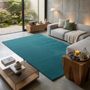 A solid teal area rug lays flat on a light grey stone floor in a modern, minimalist room. The vibrant blue-green hand-tufted rug anchors the space next to a natural wood side table with smooth black stones and a candle. A sleek beige daybed sits nearby, looking out onto a serene Japanese zen garden through large glass doors. IMG_20260727_232441.jpg displays its rich, plush texture beautifully.
