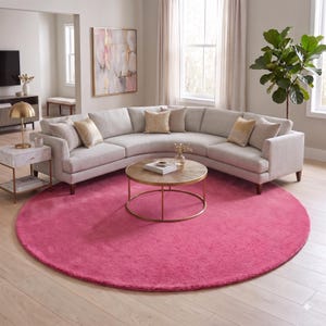 May include: A large, circular, solid hot pink area rug with a plush, textured surface. The rug is positioned on a light-colored wooden floor, serving as a vibrant foundation for a curved light gray sectional sofa and a round wooden coffee table.