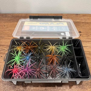 May include: A clear plastic Plano tackle box with a hinged lid containing an assortment of multi-colored, spiky, rubber fishing lures. The lures feature various shades including green, pink, blue, brown, and black, organized in compartments.