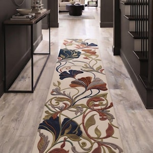 Può includere: Un lungo tappeto da corridoio rettangolare con sfondo color crema e un motivo floreale e a viticci in rilievo multicolore. Il design presenta grandi foglie e fiori stilizzati nelle tonalità del blu scuro, arancione ruggine e verde salvia.