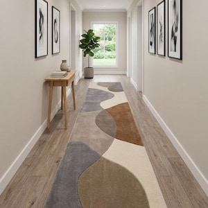 May include: A long runner rug featuring an abstract pattern of large, organic, curved shapes in shades of cream, beige, taupe, brown, and charcoal gray. The rug has a soft, plush texture and is positioned centrally along a wooden hallway floor.