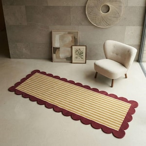 Peut inclure: Un tapis rectangulaire à rayures fines jaunes et blanches, doté d'une bordure festonnée de couleur rouge bordeaux profond. Le tapis est posé sur un sol clair dans une pièce contenant un fauteuil de couleur crème.