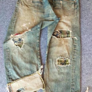 May include: Light blue denim jeans featuring a distressed finish with frayed holes and patches of camouflage fabric visible through the tears. The jeans have a button fly, rolled cuffs at the bottom, and a worn, faded appearance throughout.