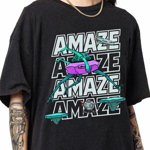 May include: A black t-shirt featuring a graphic print of the word AMAZE repeated four times in white block letters. The design includes a purple and teal insect-like creature, a small spaceship, a round robot, and two floating teal platforms.