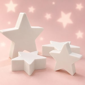May include: A set of four white star-shaped objects of varying sizes arranged together. Each star has five points and a smooth, solid surface. The objects are positioned on a light pink surface with soft, out-of-focus star shapes in the background.
