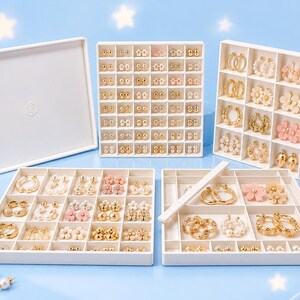 May include: A white multi-compartment jewellery organiser set featuring a lid and several trays filled with various gold-toned, pearl, and floral-shaped stud and hoop earrings. The modular storage system is designed to hold numerous pairs of earrings.