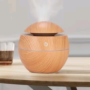 May include: A spherical essential oil diffuser with a light brown wood grain finish. The device features a removable top lid and a central power button icon on the front. A visible stream of white mist rises from the top of the diffuser.
