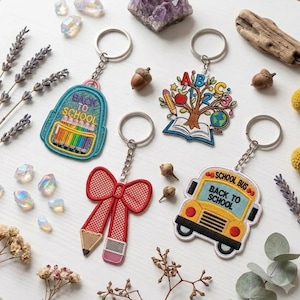 May include: Four embroidered keychains featuring school-themed designs: a teal backpack with "BACK TO SCHOOL" text, a tree growing from an open book with "ABC 123" letters, a red bow with a pencil, and a yellow school bus with "SCHOOL BUS BACK TO SCHOOL" text.