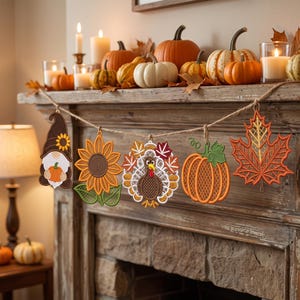 Op de afbeelding: Een decoratieve slinger met vijf geborduurde hangende ornamenten aan een touw van jute. De ornamenten bestaan uit een kabouter met een pompoen, een zonnebloem, een kalkoen omringd door herfstbladeren, een pompoen en een esdoornblad.