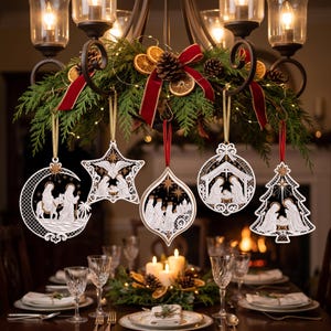 Op de afbeelding: Vijf witte, lasergesneden houten ornamenten hangen aan een kroonluchter. De ornamenten tonen gedetailleerde kersttaferelen, waaronder de vlucht naar Egypte, de ster, de drie wijzen, de kribbe en de heilige familie in een kerstboomvorm.