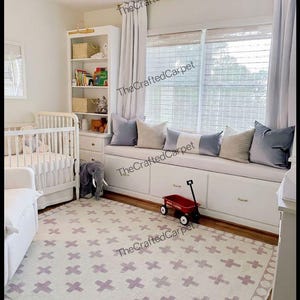 Puede incluir: Una alfombra rectangular con un patrón repetitivo de cruces de color morado claro sobre un fondo crema con un diseño de borde circular. La alfombra se encuentra en una habitación infantil con una cuna blanca, un banco bajo ventana y un carrito rojo.