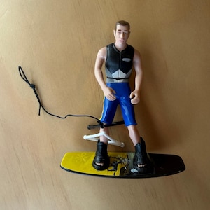 Op de afbeelding: Een plastic figuurtje van een persoon aan het wakeboarden, gekleed in een zwart zwemvest en een blauwe korte broek. Het figuurtje staat op een geel en zwart wakeboard met zwarte bindingen en houdt een wit handvat vast aan een zwart koord.