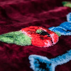 Puede incluir: Una tela de terciopelo color burdeos profundo con bordados texturizados en relieve. El dise&ntilde;o incluye una flor roja vibrante con centro negro, hojas verde claro y l&iacute;neas decorativas azul claro que se entrelazan sobre la superficie.