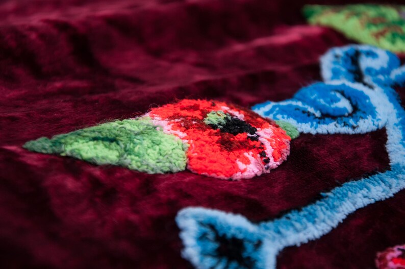 Puede incluir: Una tela de terciopelo color burdeos profundo con bordados texturizados en relieve. El dise&ntilde;o incluye una flor roja vibrante con centro negro, hojas verde claro y l&iacute;neas decorativas azul claro que se entrelazan sobre la superficie.