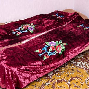 Puede incluir: Una manta de terciopelo color burdeos intenso con un destacado patr&oacute;n de bordado floral con flores rojas, moradas y verdes sobre tallos azules. La tela tiene una textura arrugada y los bordes est&aacute;n rematados con un detalle de flecos.