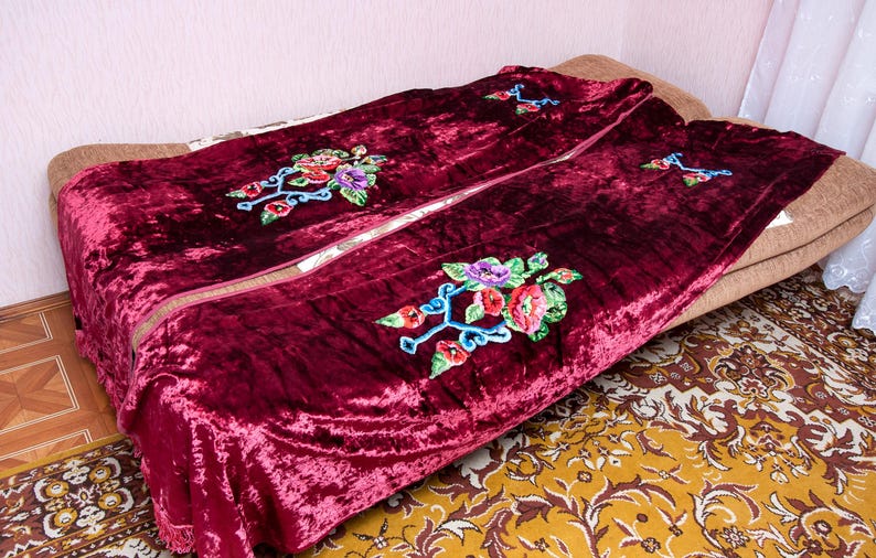 Puede incluir: Una manta de terciopelo color burdeos intenso con un destacado patr&oacute;n de bordado floral con flores rojas, moradas y verdes sobre tallos azules. La tela tiene una textura arrugada y los bordes est&aacute;n rematados con un detalle de flecos.