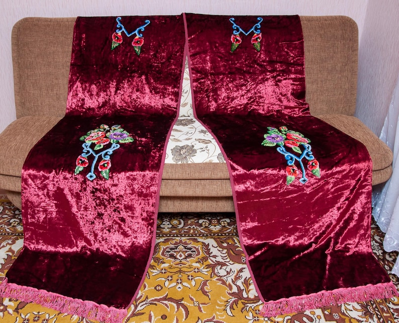 Puede incluir: Dos caminos de mesa largos de terciopelo color burdeos intenso con intrincados bordados florales en tonos rojo, morado y verde, acentuados con volutas azul claro. Cada camino tiene un borde de flecos a juego en la parte inferior.