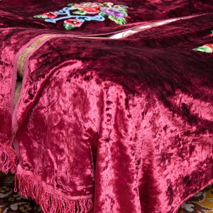 Puede incluir: Una manta de terciopelo arrugado en color burdeos intenso con flecos decorativos en los bordes y motivos florales bordados en tonos vibrantes de rojo, azul, verde y amarillo, mostrando una superficie de tela texturizada con brillo.