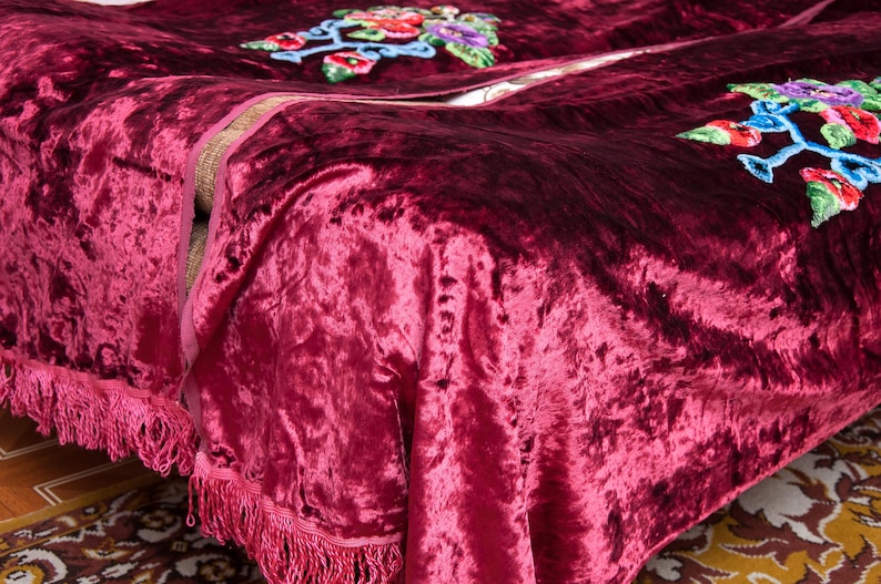 Puede incluir: Una manta de terciopelo arrugado en color burdeos intenso con flecos decorativos en los bordes y motivos florales bordados en tonos vibrantes de rojo, azul, verde y amarillo, mostrando una superficie de tela texturizada con brillo.