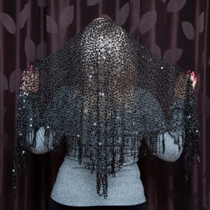 May include: A black sequined shawl with fringe. The shawl is draped over a person, obscuring their face. The sequins are densely packed, creating a shimmering effect. The fringe hangs from the bottom edge of the shawl.
