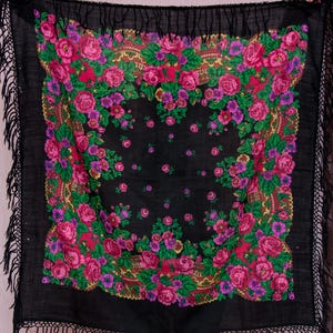 Op de afbeelding: Een zwarte sjaal met een bloemenpatroon van roze, paarse en groene rozen. De sjaal heeft een franjerand en een decoratieve rand met gouden accenten. Het ontwerp is symmetrisch.