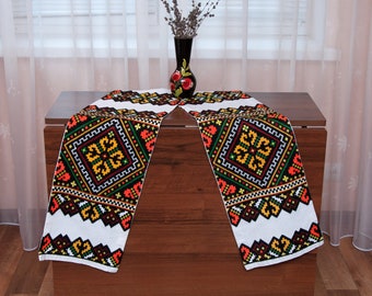 May include: Two white table runners with black, red, yellow, and green embroidered geometric patterns. The runners are laid out on a wooden table with a vase of dried lavender in the center.