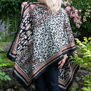Op de afbeelding: Een vierkante poncho met een luipaardprint in zwart, wit en perzik. De poncho heeft een zwarte rand met een perzikstreep en een decoratief patroon. Het kledingstuk is over de schouders gedrapeerd.