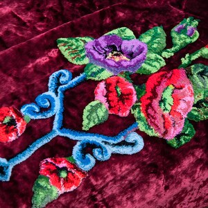 Puede incluir: Un tejido de terciopelo color burdeos intenso con un dise&ntilde;o central de bordado con aguja m&aacute;gica. El bordado muestra flores vibrantes en rojo, rosa y morado con hojas verdes, acentuadas por patrones decorativos de volutas azul brillante.