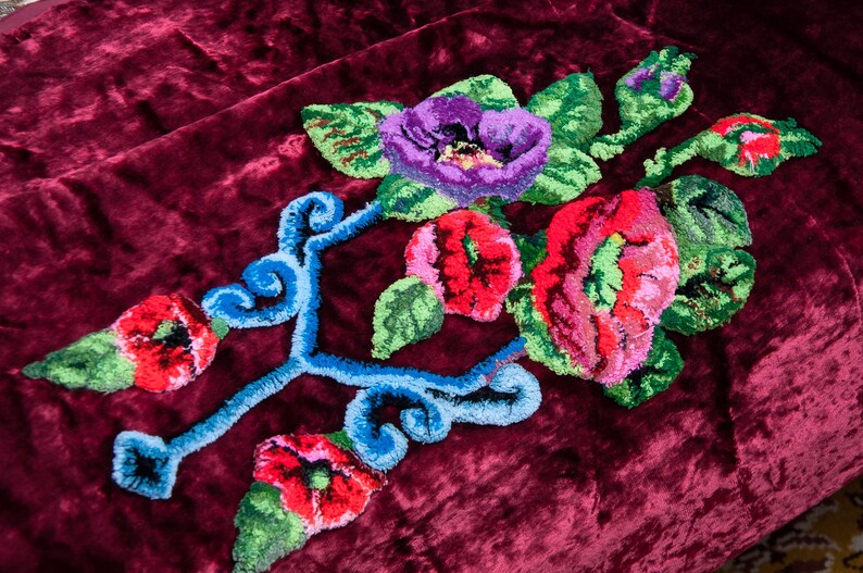 Puede incluir: Un tejido de terciopelo color burdeos intenso con un dise&ntilde;o central de bordado con aguja m&aacute;gica. El bordado muestra flores vibrantes en rojo, rosa y morado con hojas verdes, acentuadas por patrones decorativos de volutas azul brillante.