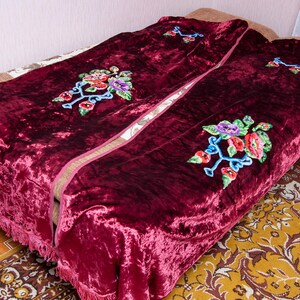 Puede incluir: Una manta de terciopelo color burdeos intenso con una textura arrugada y flecos decorativos en los bordes. La tela est&aacute; adornada con motivos florales bordados en tonos rojo, morado, verde y azul, dispuestos en un patr&oacute;n repetitivo.