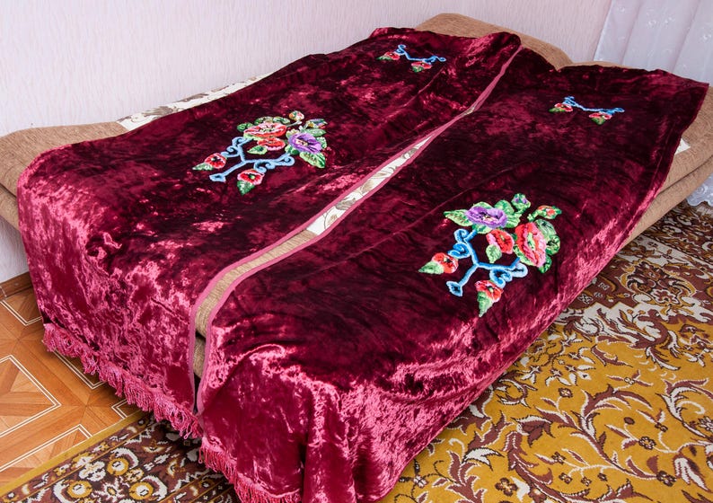 Puede incluir: Una manta de terciopelo color burdeos intenso con una textura arrugada y flecos decorativos en los bordes. La tela est&aacute; adornada con motivos florales bordados en tonos rojo, morado, verde y azul, dispuestos en un patr&oacute;n repetitivo.