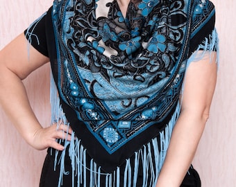 May include: A black, blue, and white patterned scarf with fringe. The scarf features a floral and paisley design in shades of blue, black, and white. The scarf is draped over the shoulders.