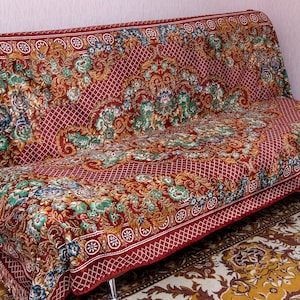 May include: A sofa covered in a deep red fabric featuring an intricate, multi-colored floral pattern with green, gold, and blue accents. The textile has a lattice design, a decorative border with circular motifs, and fringe along the edges.