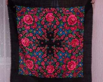 May include: A square black shawl with a vibrant floral pattern. The design features large pink roses, blue and yellow flowers, and green leaves. The shawl has a fringed edge and is held up by unseen hands.