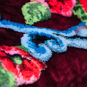 Puede incluir: Un tejido de terciopelo color burdeos profundo con bordados en relieve de colores vibrantes. El dise&ntilde;o incluye flores de color rojo brillante con hojas verdes y patrones de enredaderas en azul claro, todo con un acabado de hilo grueso.