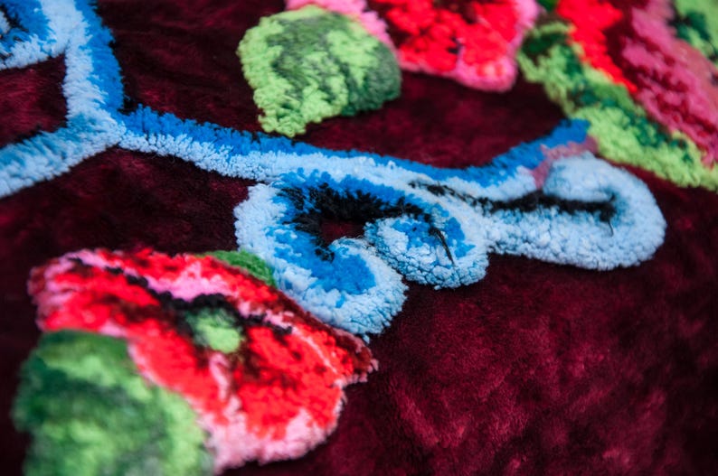 Puede incluir: Un tejido de terciopelo color burdeos profundo con bordados en relieve de colores vibrantes. El dise&ntilde;o incluye flores de color rojo brillante con hojas verdes y patrones de enredaderas en azul claro, todo con un acabado de hilo grueso.