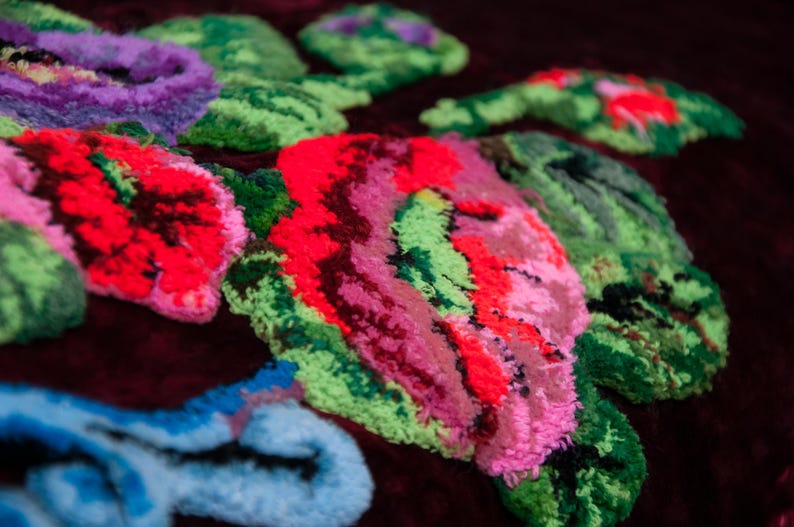 Puede incluir: Una tela de terciopelo color burdeos oscuro con un patr&oacute;n de bordado floral en relieve y texturizado. El dise&ntilde;o incluye flores grandes y vibrantes en tonos rosa ne&oacute;n, rojo brillante, verde lima y azul claro, hechas con hilo grueso.