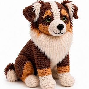 Op de afbeelding: Een gehaakte knuffelhond met een driekleurige vacht van donkerbruin, beige en crèmekleurig garen. Het pluche dier heeft groene plastic ogen, een zwart geborduurde neus en mond, en een pluizige crèmekleurige borstkraag met bijpassende oorpluimen.