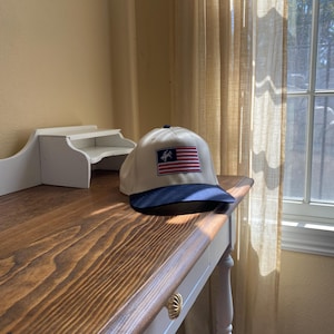 May include: A baseball cap with a cream-coloured crown and a dark blue brim. The front panel features a rectangular patch displaying a stylised American flag with a small silhouette of a bucking horse and rider positioned over the blue field.